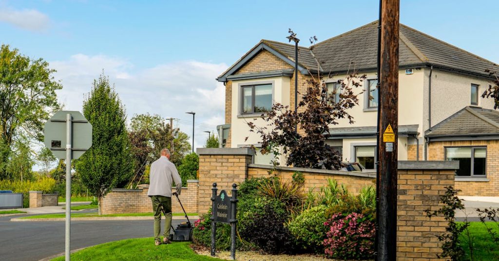 A man mowing the lawn outside a modern suburban home with a lush garden and clear blue sky.