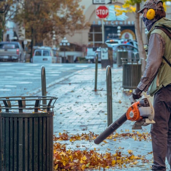 Street worker using a leaf blower during autumn in a city setting, surrounded by fallen leaves.
